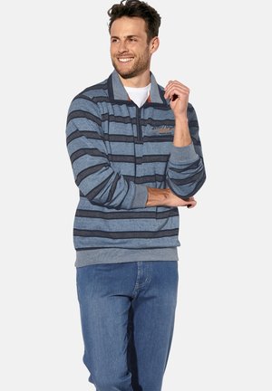 Ein Mann lächelt, trägt einen blau gestreiften Pullover mit Viertel-Reißverschluss und blauen Jeans. Er steht mit einem Arm verschränkt und die andere Hand nah an seinem Gesicht.