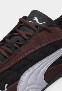 Κοντινό πλάνο ενός μαύρου και καφέ αθλητικού παπουτσιού με λευκό λογότυπο Puma και καφέ κορδόνια σε ανοιχτό φόντο.