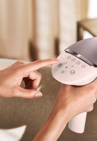 Appareil manuel avec un boîtier blanc arrondi et un dessus gris. Présente un panneau de commande avec des boutons et un doigt appuyant sur un bouton d'alimentation.