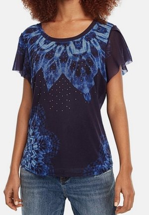 Kobieta ubrana w granatową bluzkę z niebieskimi wzorami tie-dye oraz przezroczystymi zwiewnymi rękawami, zestawioną z niebieskimi dżinsami.