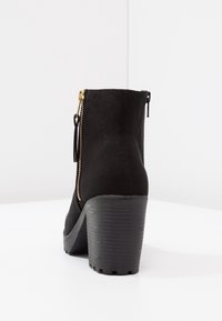 Czarne zamszowe botki za kostkę na wysokim, masywnym obcasie, z ozdobnym złotym zamkiem po boku oraz teksturowaną gumową podeszwą. Prosty i elegancki design.