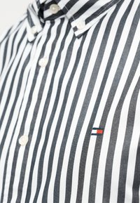 Πουκάμισο με κουμπιά και κάθετες μαύρες και λευκές ρίγες, με ένα μικρό λογότυπο σε κόκκινο, λευκό και μπλε στο αριστερό μέρος του στήθους. Υλικό από βαμβάκι.