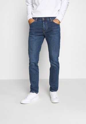 Man draagt slim-fit blauwe jeans en witte sneakers met handen in de zakken tegen een effen witte achtergrond.