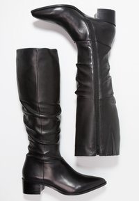 Botas de cuero negro hasta la rodilla con punta afilada, tacón bloque moderado y cremallera lateral; presentan un diseño gather a lo largo del eje.