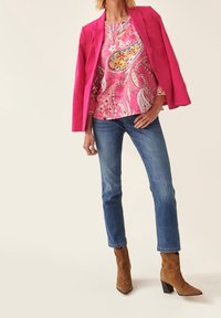 Blazer rose sur un haut à motif paisley, associé à un jean bleu et des bottines à talons en suède marron. Fond clair.