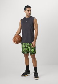 Μαύρη αμάνικη μπλούζα, πράσινα σορτς με σχέδιο φίδι, μαύρα αθλητικά με πορτοκαλί λεπτομέρειες, κρατώντας ένα μπάσκετ, σε ουδέτερο φόντο.