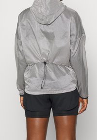 Šedý kostkovaný větrový kabát s kapucí, staženým pasem, elastickými manžetami a hladkou, lehkou texturou. Nosit přes černé šortky.