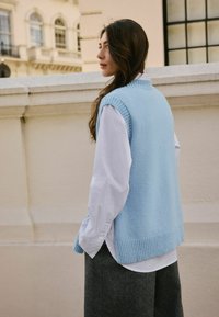 Lichtblauwe gebreide sweater met een geribbelde zoom, gedragen over een witte blouse met kraag. Donkergrijze gestructureerde broeken. Zachte, gezellige uitstraling.