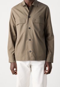 Hnědá košile s dlouhým rukávem, límečkem, dvěma náprsními kapsami a zapínáním na knoflíky. Vyrobena z hladké látky s jemnou texturou.