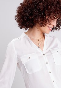 Camicia bianca con bottone in tessuto leggero, due tasche sul petto, dettagli in metallo e una texture morbida, abbinata a collane dorate a strati.