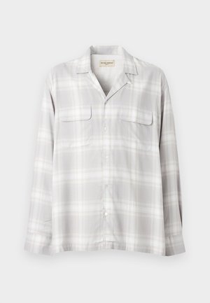 Άνδρας που φορά ανοιχτό γκρι καρό overshirt, λευκό t-shirt, σκούρο καφέ παντελόνι και μαύρα πλεκτά δερμάτινα παπούτσια χωρίς κορδόνια, στέκεται με το ένα χέρι στην τσέπη.