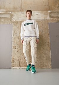 Vit sweatshirt med grön text "Originals" och marinblå ränder, beige cargo-byxor med fickor och gröna sneakers på ett slätt golv.