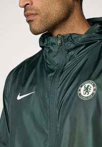 Green Nike jacket with a zippered front, hood, and Chelsea Football Club logo on the left chest. Smooth, lightweight fabric with a shiny finish.