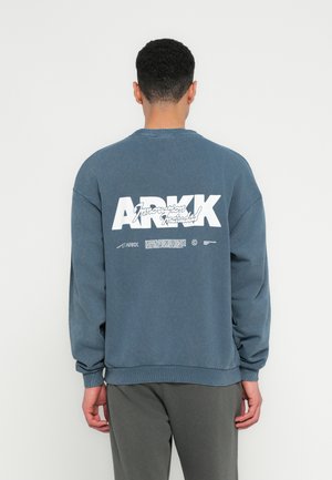 Homme portant un sweat-shirt bleu avec un grand "ARKK" blanc et un texte cursif plus petit au dos, debout face à un fond blanc uni.