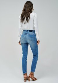 Blusa blanca ligera de manga larga, combinada con jeans de mezclilla ajustados de cintura alta y bolsillos traseros. Sandalias en tonos neutros.