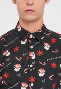 Chemise noire à boutons avec un motif de fête représentant des visages de Santa, des cannes de Noël, des flocons de neige et le texte "HOLIDAYS HEUREUX".