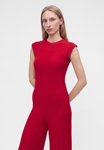 Femme aux longs cheveux blonds portant une combinaison rouge sans manches, debout les mains derrière le dos devant un fond clair uni.