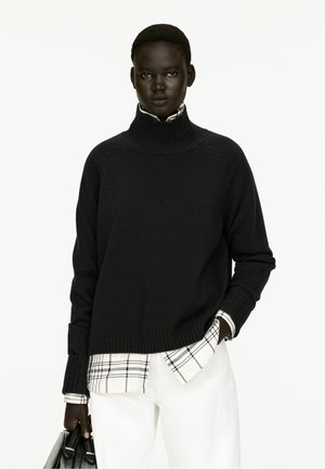 Pull noir à col roulé avec des poignets et un ourlet côtelés, superposé sur une chemise à carreaux noir et blanc, associé à un pantalon blanc, tenant un sac noir.