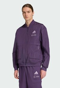 Purple lightweight bomber jacket with a zip front, two front pockets, and Adidas branding. Textured fabric and elastic cuffs and hem.