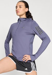 Haut de sport à manches longues violet avec un col à moitié zippé, présentant des rayures grises sur les manches et un design ajusté pour faciliter le mouvement.