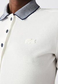 Polo shirt blanc avec un tissu texturé, doté d'un col rayé marine, de trois boutons et d'un logo brodé sur la poitrine.