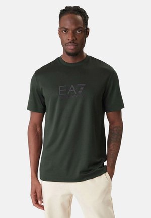 Hombre con camiseta EA7 Emporio Armani de color verde oscuro y pantalones beige claros, de pie con una mano en el bolsillo sobre un fondo liso.