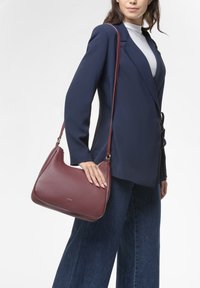 Borsa in pelle bordeaux con forma arrotondata, dotata di una lunga tracolla regolabile e di un sottile marchio dorato. Indossata sopra un blazer blu e jeans.