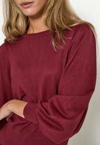 Bordeauxfarbener Strickpullover mit Rippenkragen und -bündchen, der leicht gepuffte lange Ärmel und einen strukturierten Körper aufweist.