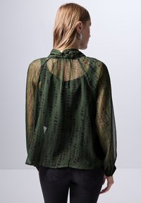 Blusa verde trasparente con maniche lunghe, caratterizzata da un delicato motivo di strisce più scure e un dettaglio al collo annodato. Jeans neri completano il look.