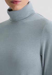 Person, die einen hellblauen Rippstrick-Turtleneck-Pullover trägt, der bis zum Kinn und oberen Brustbereich gekürzt ist, vor einem schlichten hellen Hintergrund.