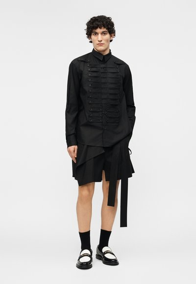 Giovane con una camicia nera abbottonata con strisce decorative, gonna asimmetrica nera, calzini neri e mocassini bianchi e neri.