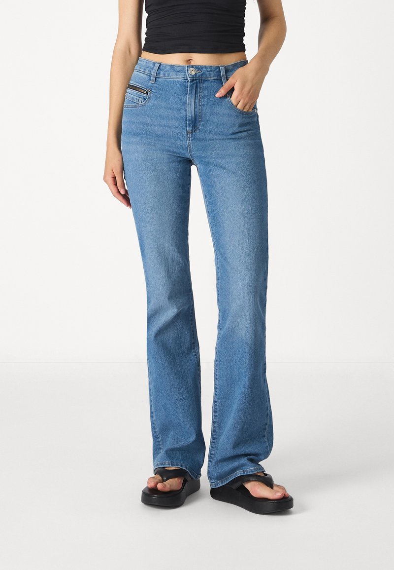 Jeans acampanados de color azul claro, hechos de mezclilla, con una cintura de altura media, bolsillos frontales y un detalle de bolsillo con cremallera, combinados con sandalias negras.