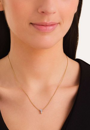 Collar de cadena de oro con un pequeño colgante en forma de letra "T" incrustado de diamantes, reposando sobre la clavícula. Diseño simple y elegante.