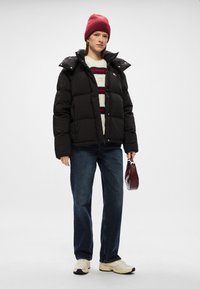 Μαύρο τζάκετ από πούπο�υλα με ψηλό γιακά, φορεμένο πάνω από ένα κρεμ και κόκκινο ρίγες πουλόβερ. Σκούρο μπλε τζιν και λευκά αθλητικά παπούτσια. Κρατάει μια μικρή μπορντό τσάντα.