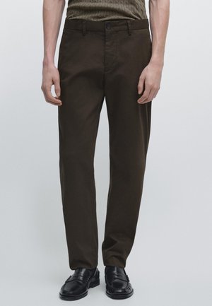 Mann trägt dunkelbraune Hose, schwarze Loafers und ein olivgrünes Strickoberteil, steht vor einem einfachen hellen Hintergrund.