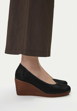 Scarpe con zeppa nere con piattaforma marrone strutturata indossate con pantaloni marroni corti su piedi nudi.