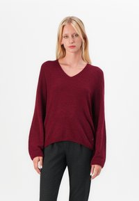 Maglione bordeaux a V con maniche lunghe, texture morbida e vestibilità comoda. Abbinato a pantaloni neri a righe, creando un contrasto di stili.