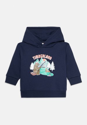 Tengerészkék kisgyermek kapucnis pulóver, medvével, amely a folyóparton horgászik, hegyekkel, fákkal és a „Timberland” szóval a jelenet fölött.