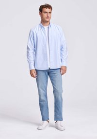 Man staat rechtop en draagt een lichtblauw overhemd over een wit T-shirt, lichtblauwe jeans en witte sneakers, tegen een effen lichte achtergrond.