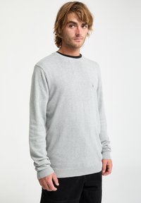 Jasnoszary sweter z miękkiego materiału, z okrągłym dekoltem i prążkowanymi mankietami; mały detal logo na klatce piersiowej, w połączeniu z czarnymi spodniami.