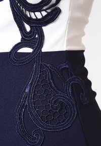 Námořnické modré šaty s jemnými bílými vyšívanými spirálovými vzory a hexagonálními motivy, které zdůrazňují kontrastní textury a propracované řemeslné zpracování.