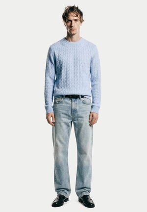 Junger Mann steht, trägt hellblauen Zopfmusterpullover, ausgewaschene blaue Jeans mit schwarzem Gürtel und schwarze Schuhe, vor schlichtem weißem Hintergrund.