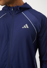 Tumši zils sporta žakets ar kapuci, ar gaiši pelēkiem akcentiem, ar rāvējslēdzēja priekšpusi un teksturētu paneli uz pleca. Rotāts ar logotipu.