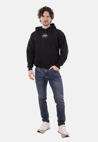 Felpa nera con tasca anteriore, coulisse regolabile e design grafico. Abbinata a jeans blu e sneaker bianche.