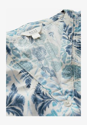 Camicia di lino con motivo floreale in blu e verde chiaro su sfondo bianco. Presenta una chiusura con bottoni e una texture morbida e leggera.