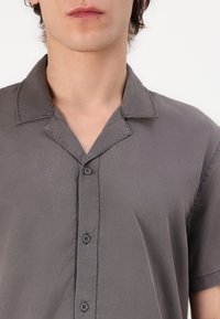 Chemise grise à manches courtes en tissu lisse. Présente un col à encoches et une fermeture à boutons sur le devant avec des boutons foncés. Design épuré et minimaliste.