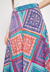 Žena v barevném patchworkovém sukni s květinovými a geometrickými vzory v odstínech modré, zelené, oranžové, růžové a fialové, v kombinaci s bílým topem.