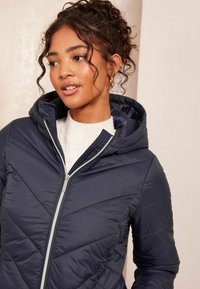 Jūras spēku pufīgs jaku ar kapuci, ar ševronu kvadrātveida struktūru un sudraba rāvējslēdzēju, uzvilkts pār krēmkrāsas ribota svītera.