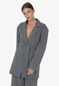 Blazer grigio a righe con vita con cintura, rever a punta e vestibilità ampia. Presenta maniche lunghe e tasche laterali applicate.