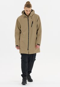 Beige wasserdichter langer Mantel mit einem schwarzen Reißverschluss und einer Brusttasche, kombiniert mit schwarzen Cargo-Hosen und schwarzen Wanderschuhen. Glatte Textur.